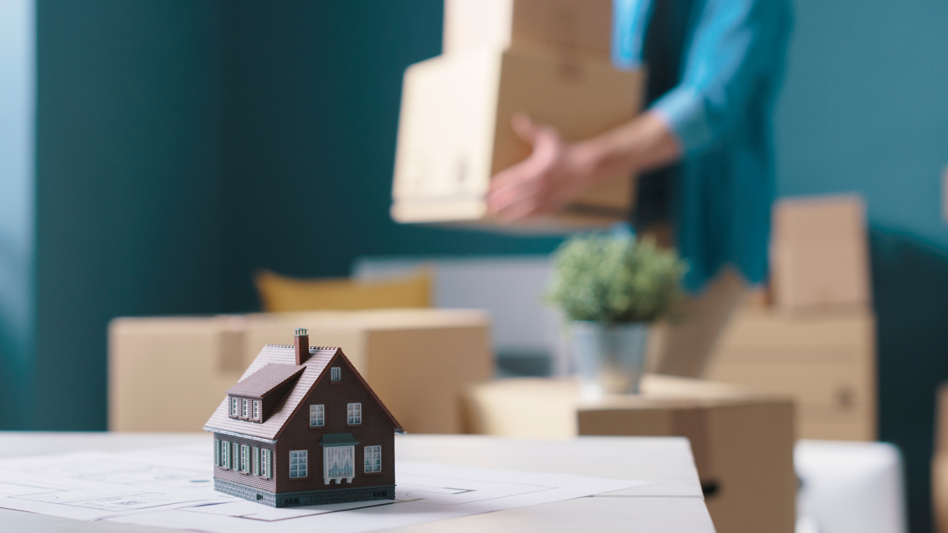 Miniature house on table; person carries moving boxes in new home.