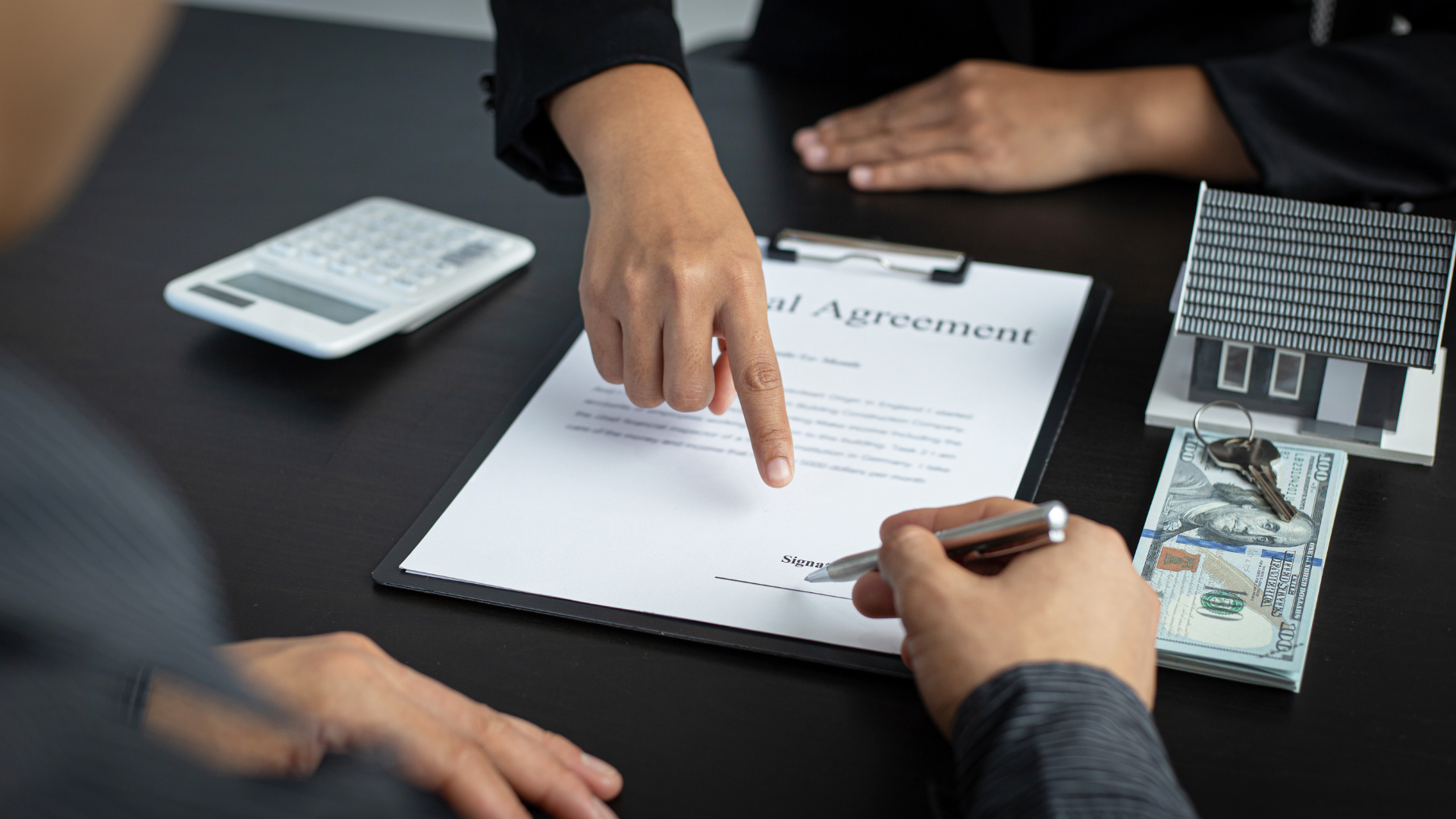Person signing a real estate agreement, another pointing at a line; calculator and keys visible.