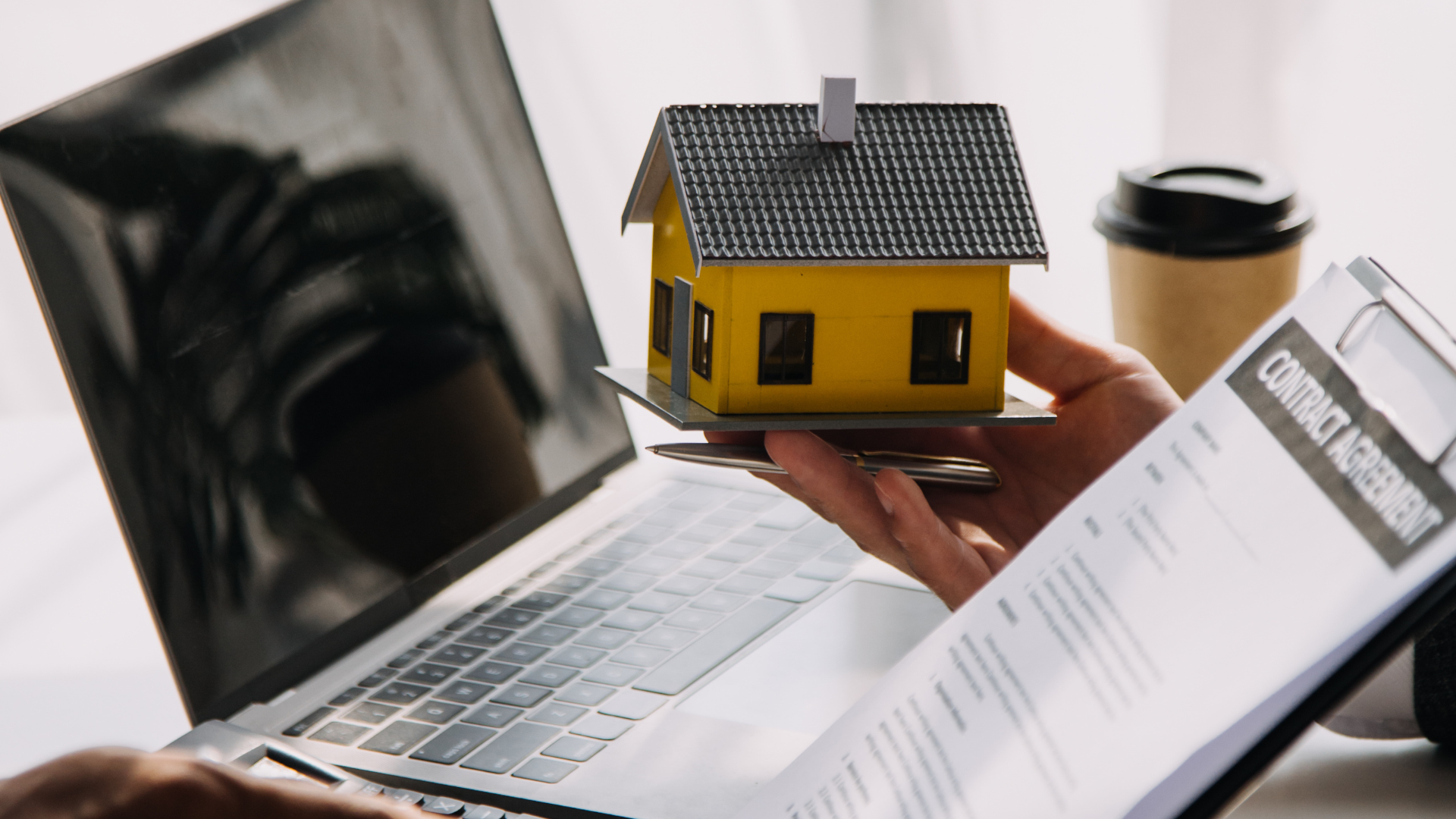 Person holding model house, laptop, paperwork, and coffee cup; real estate concept.