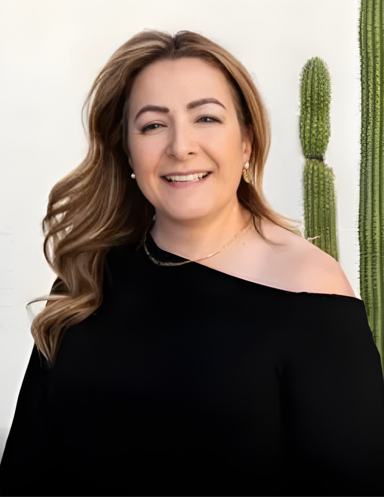 Woman with long wavy blonde hair smiles, wearing a black off-the-shoulder top, standing in front of a cactus.