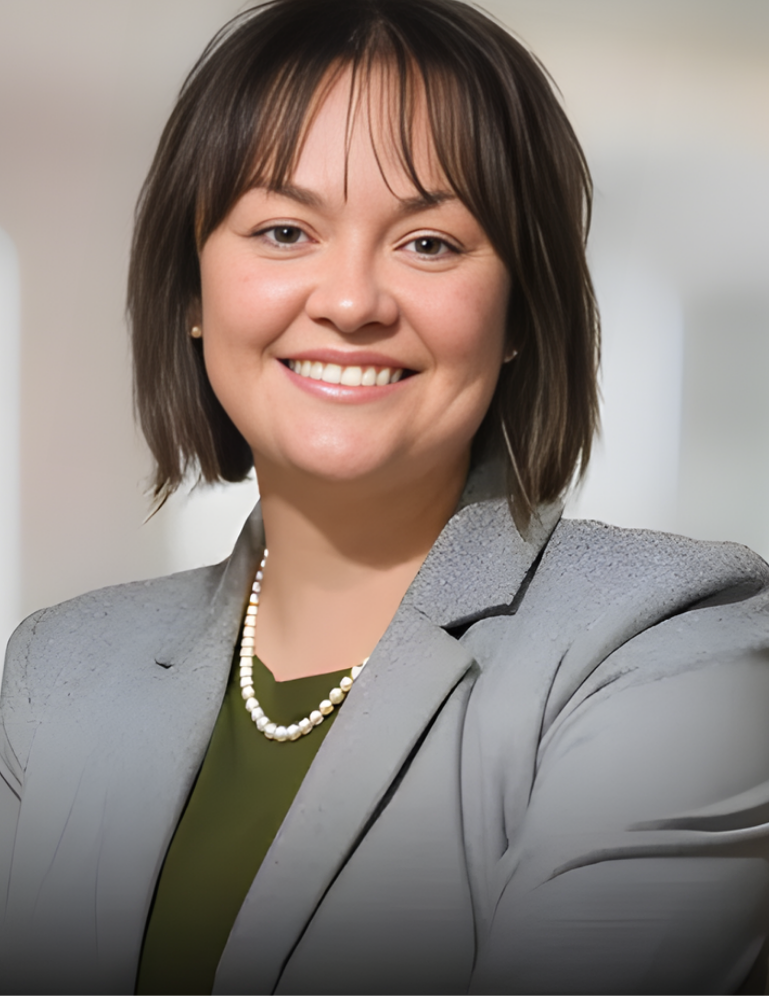 Woman in a grey blazer and pearl necklace smiles, with a green top. Blurred background.