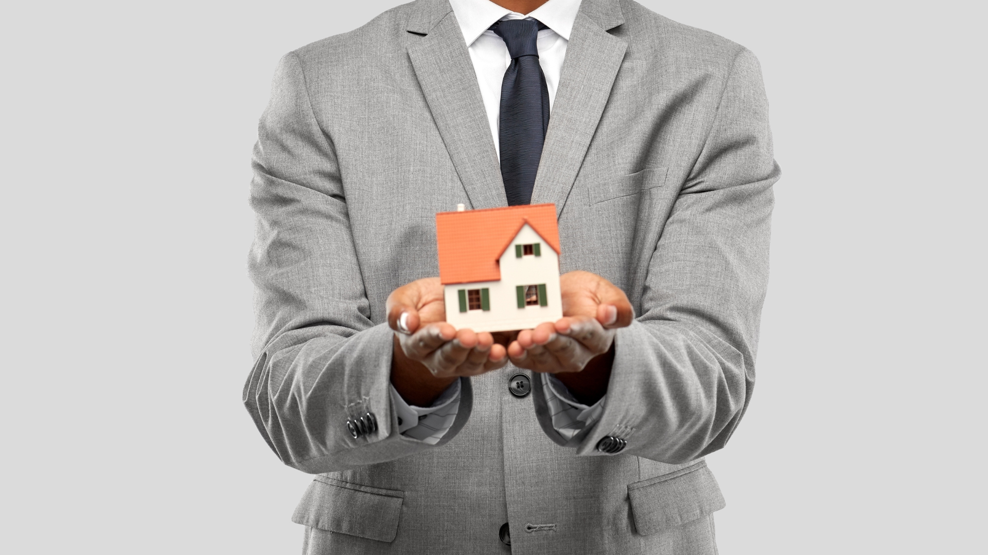 Person in gray suit holding a miniature house, possibly real estate concept.