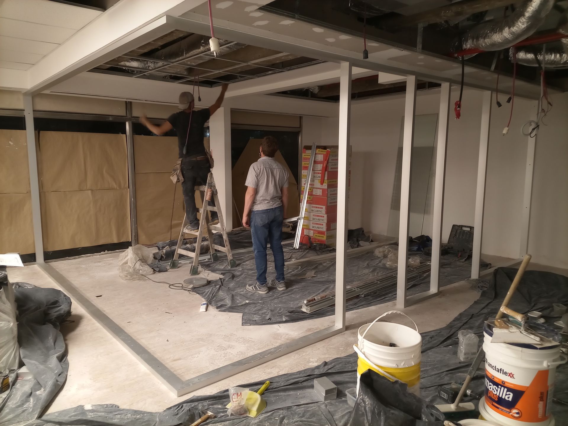 Dos personas construyendo una pared interior; materiales de construcción y herramientas visibles.