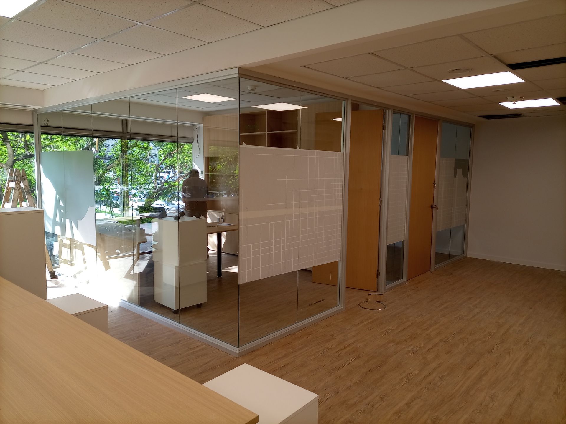 Interior de oficina con paredes de vidrio, pisos de madera y una persona trabajando en un escritorio