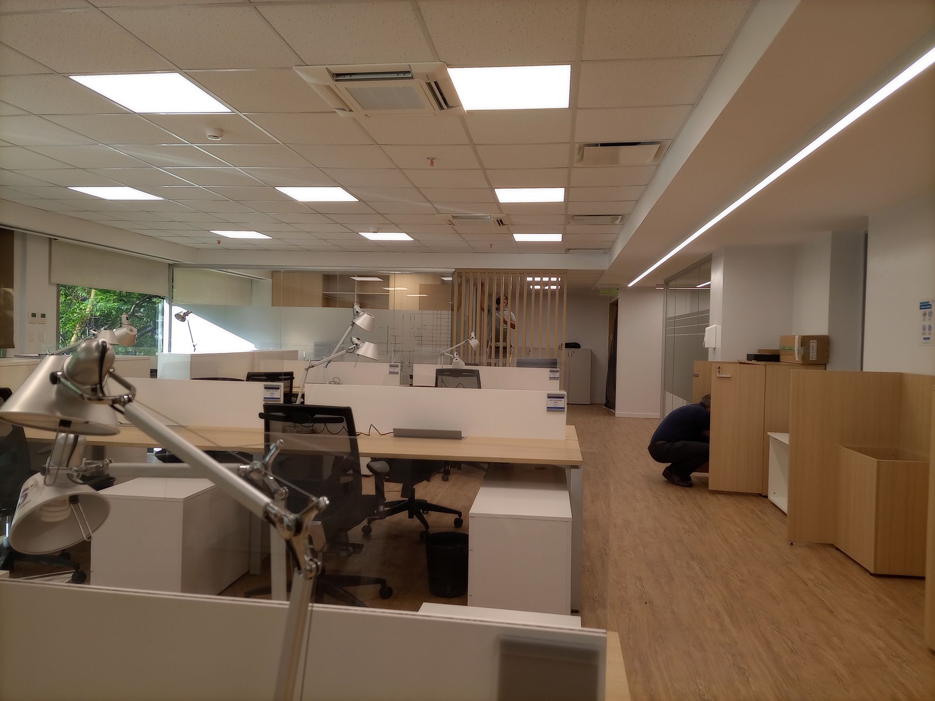 Interior de oficina con escritorios, sillas e iluminación cenital; una persona se agacha junto a cajas de cartón.