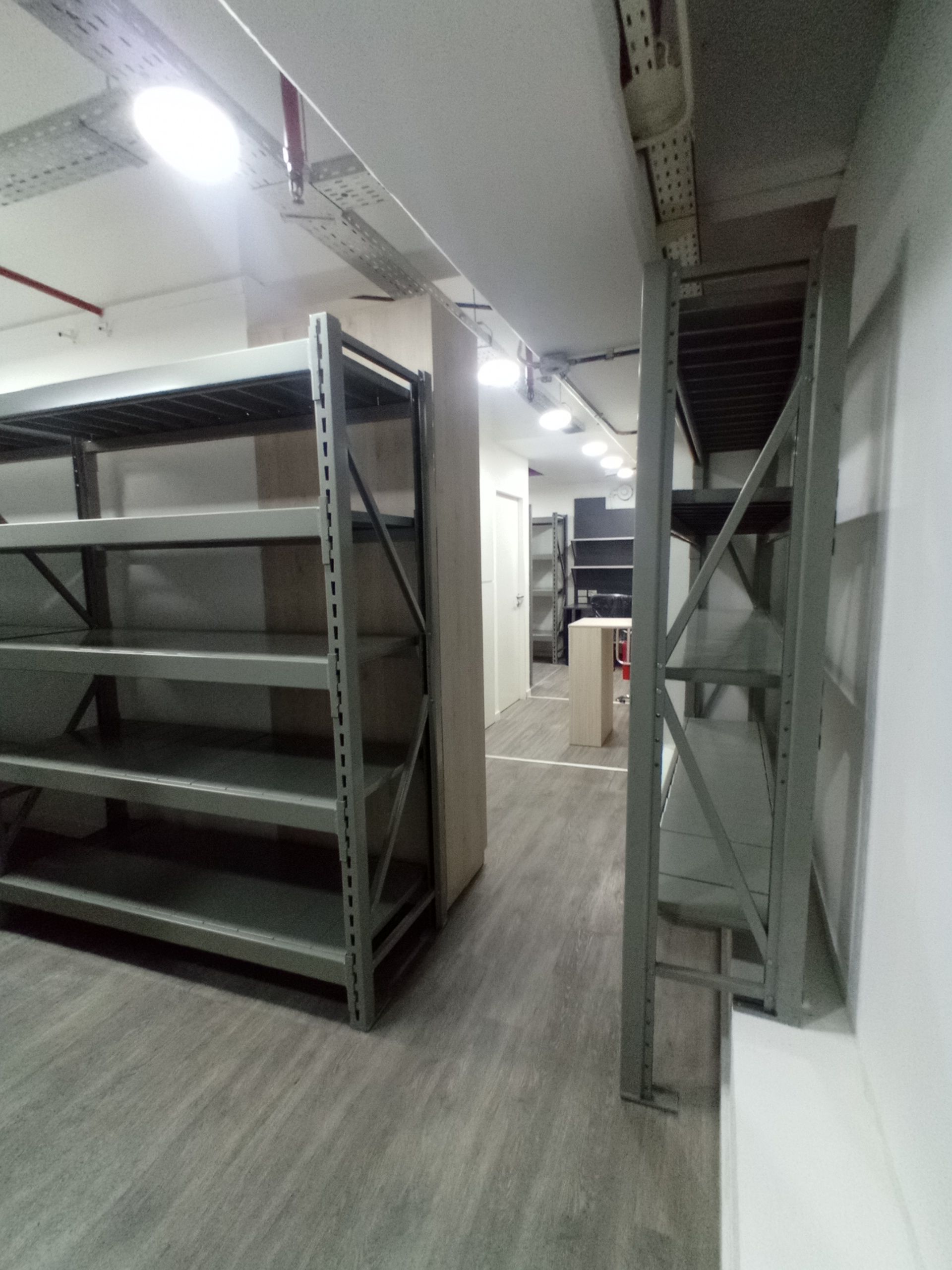 Estantes metálicos vacíos en un trastero con suelo gris, pared blanca y techo bajo con luces superiores.