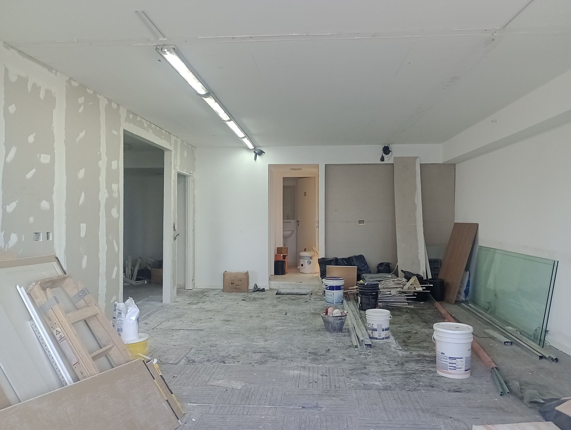 Espacio interior en construcción; paneles de yeso y materiales esparcidos en el piso; iluminación cenital.