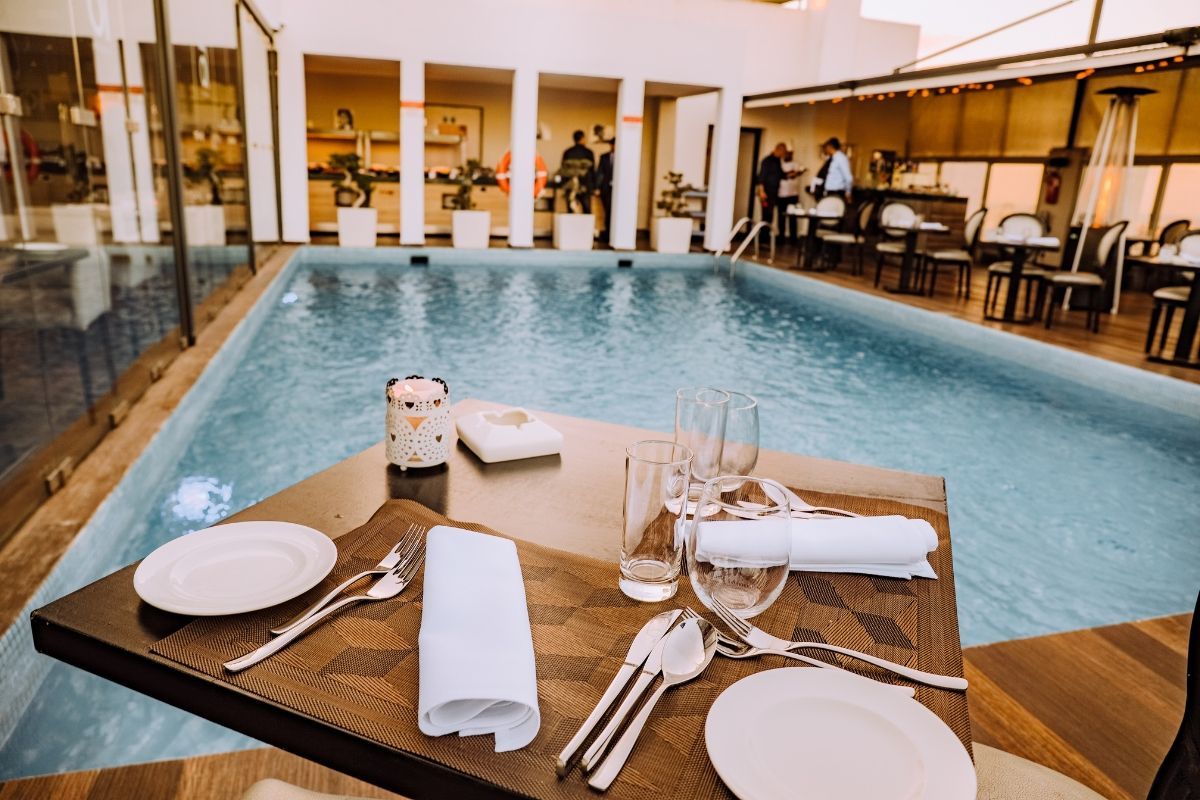Mesa de restaurante posta ao lado de uma piscina coberta, com pratos brancos e guardanapos dobrados.