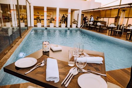 Elegante conjunto de mesa e cadeiras para refeições à beira da piscina, com pratos, copos e guardanapo enrolado, sobre a água.