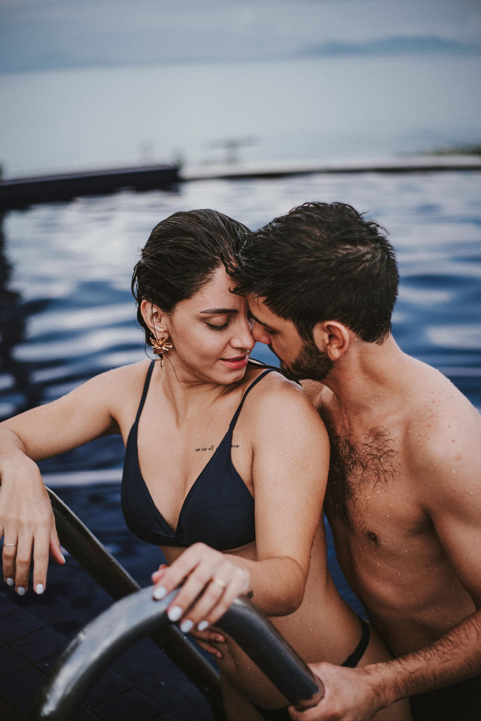 Duas pessoas estão sentadas próximas uma da outra em um barco, sorrindo sobre a água com a linha costeira desfocada ao fundo.