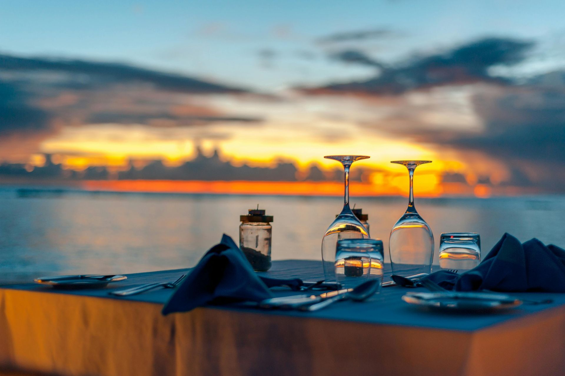 Jantar ao pôr do sol à beira-mar com taças de vinho, pratos, guardanapos e um horizonte desfocado ao fundo.