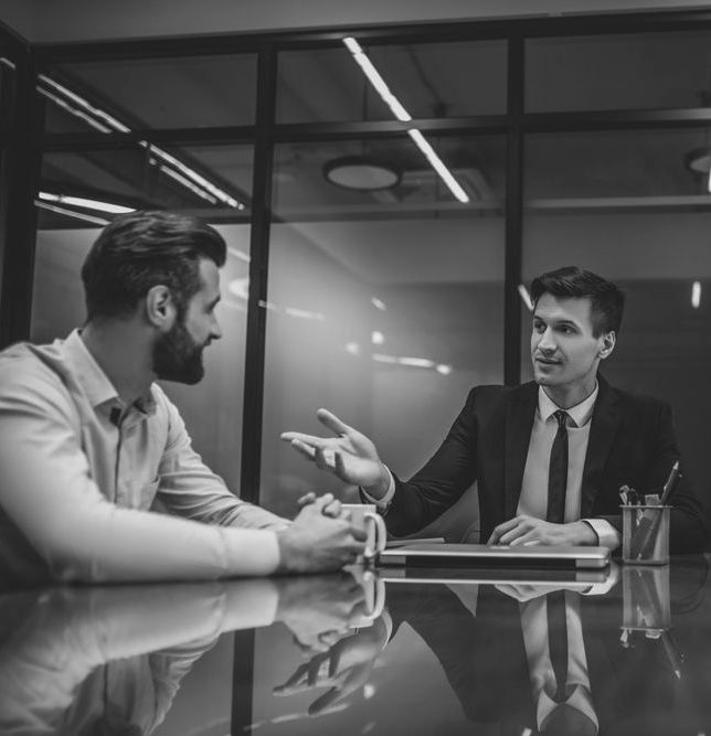 Two men in a business meeting, one speaking across a conference table in a glass-walled office. - ev law in Bundall, QLD