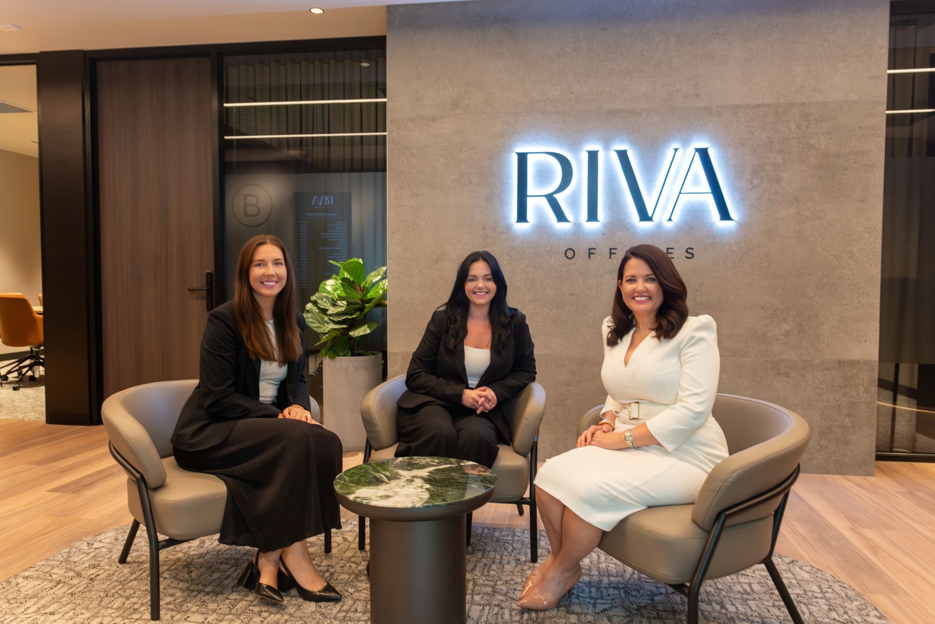 Three people sitting in chairs around a small table in front of a wall with a backlit 