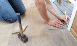 Person kneeling, measuring flooring with a pencil, hammer nearby.