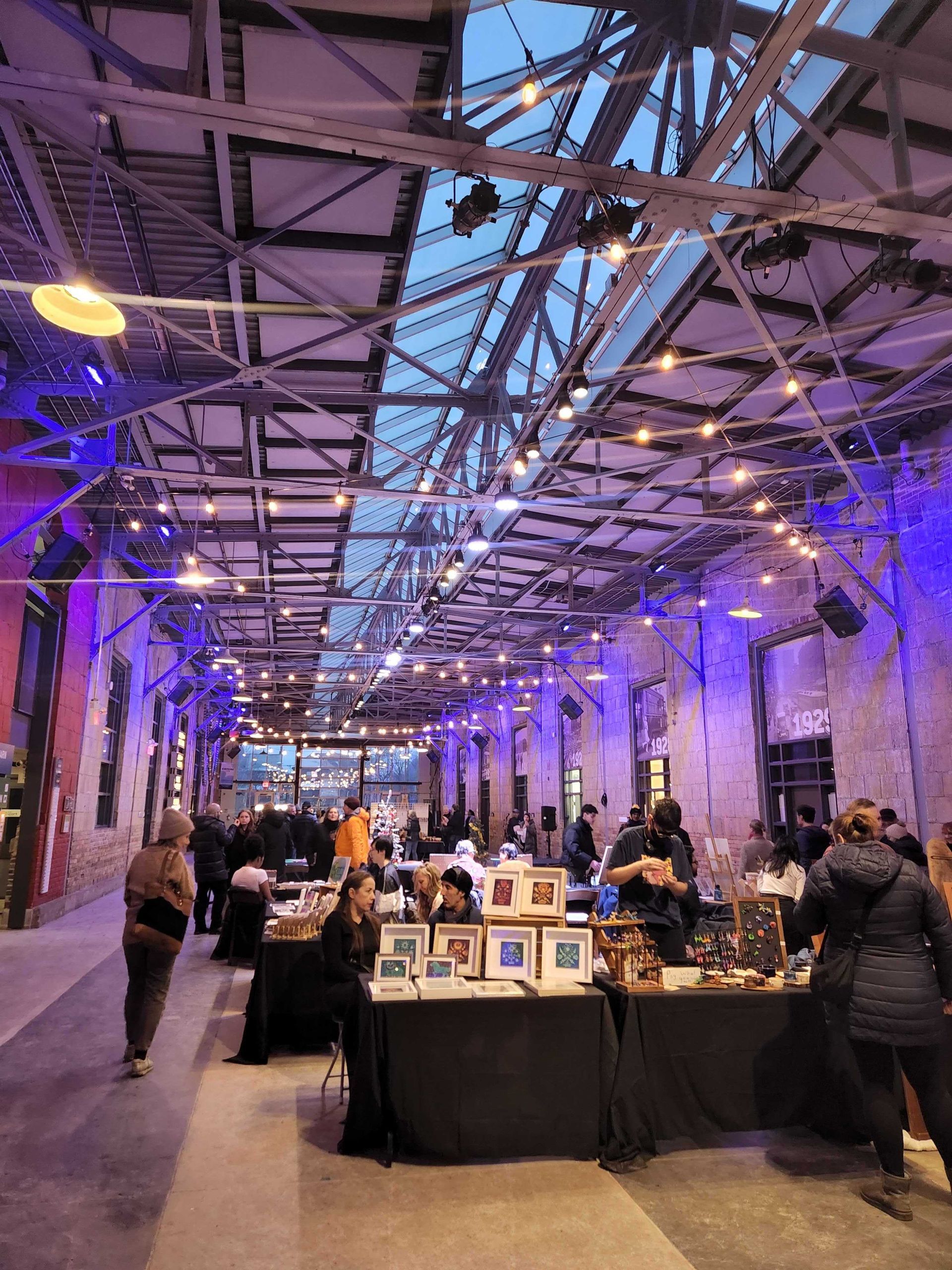 Indoor market with vendors and shoppers under a high-ceilinged industrial building. Blue and purple lights.