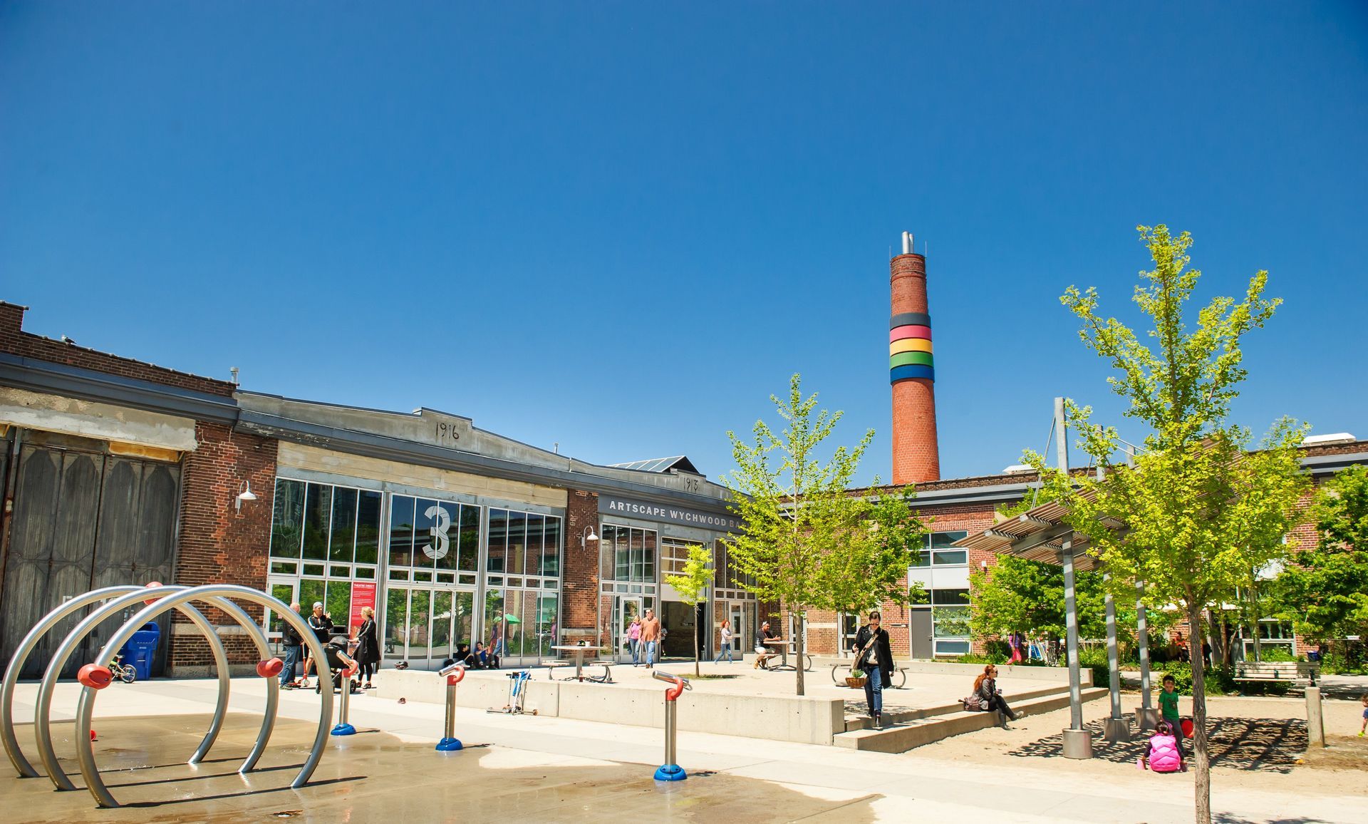 Daytime view of a brick building with a playground, trees, and a colorful chimney under a blue sky.