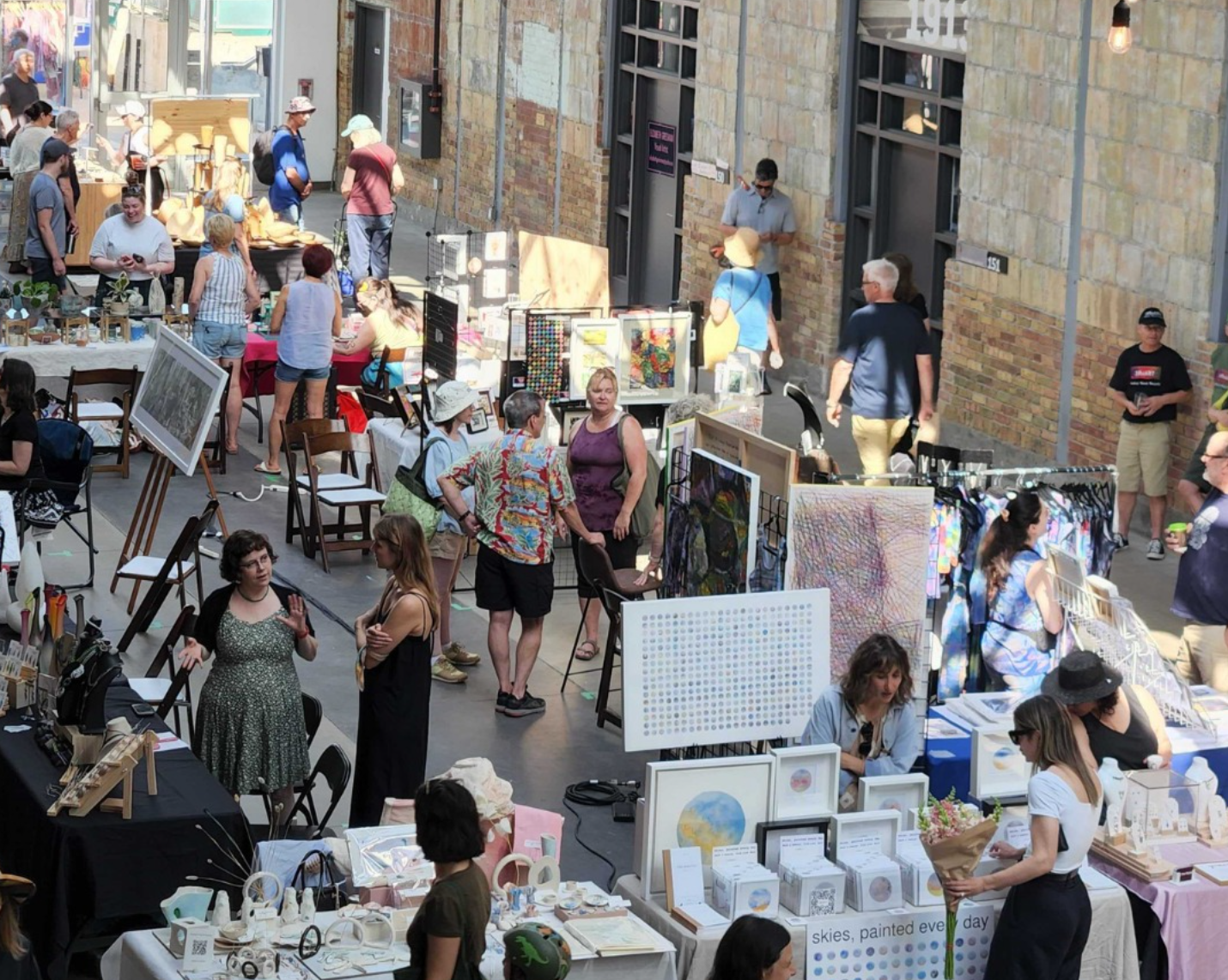 People browsing art and crafts at an indoor market, diverse vendors and shoppers, sunny lighting.