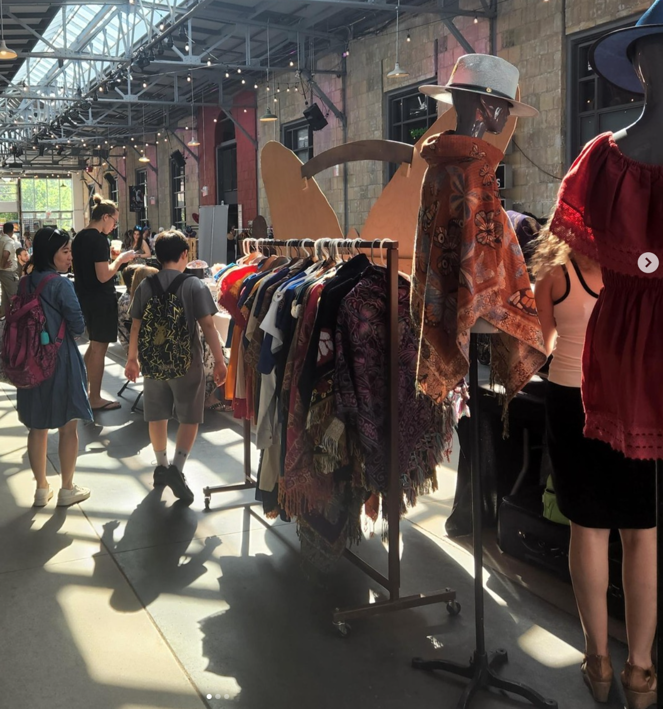 Clothing rack at an outdoor market; people browse, sunlight streams in from above.