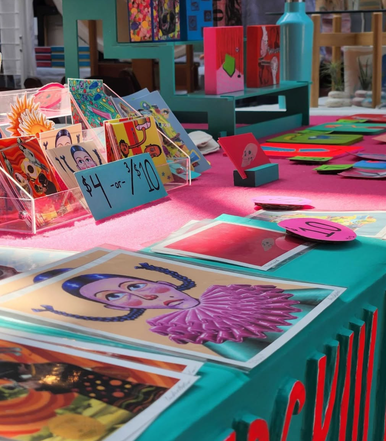 Art display table with colorful artwork on a pink tablecloth.