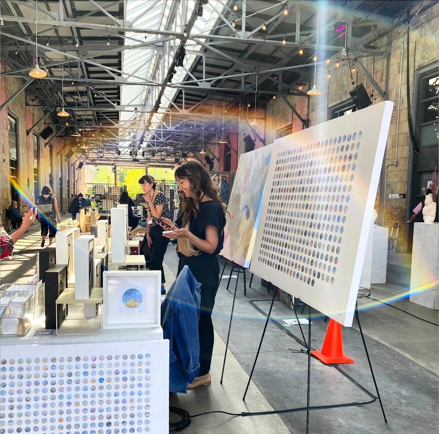 Art show: woman looks at a large white painting on an easel. Others view art in a large, open industrial space.