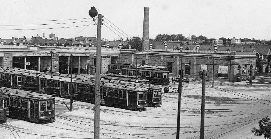 Streetcars parked in a yard in front of multiple garages; power lines and a tall smokestack in the background.