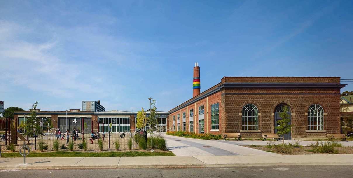 Brick industrial building with smokestack and modern glass structure under blue sky.