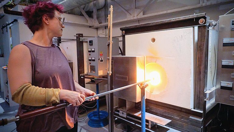 Woman with pink hair in a workshop, holding a metal rod near a glowing furnace.