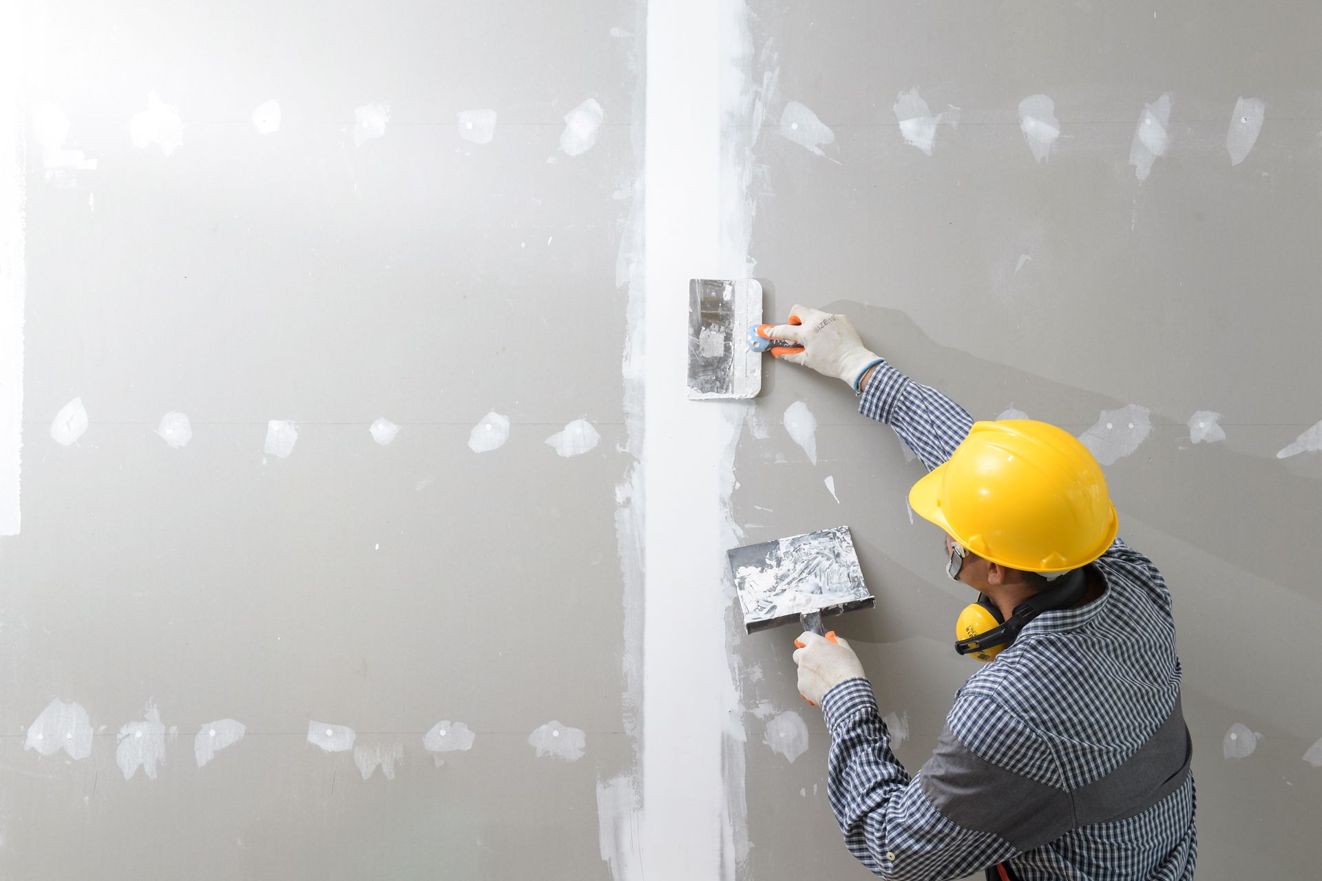 Person in yellow hard hat applying drywall compound to a wall with two putty knives.