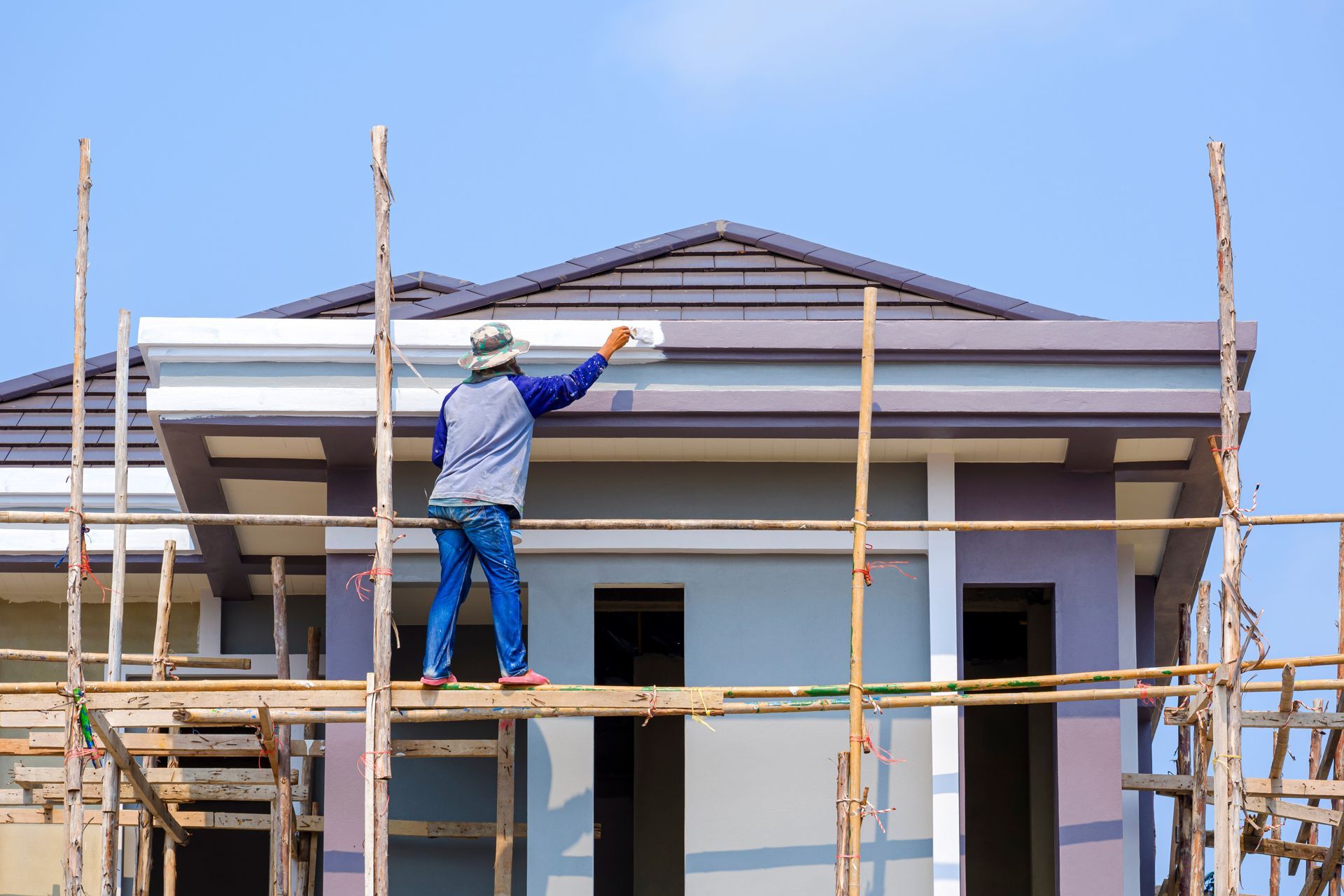 Construction worker painting a house's trim while standing on scaffolding against a blue sky.