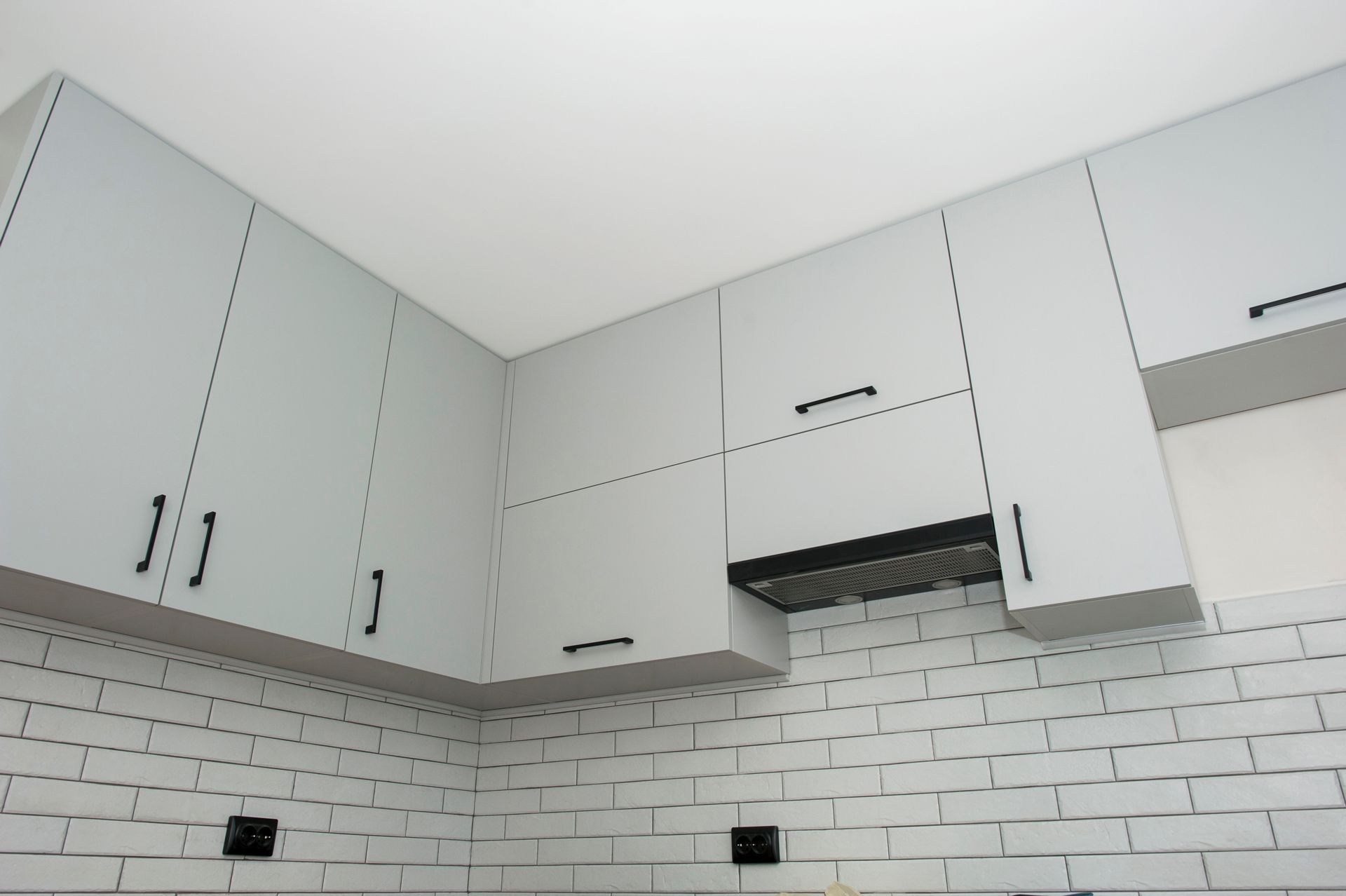 Gray kitchen cabinets with black handles above a white brick backsplash and range hood.