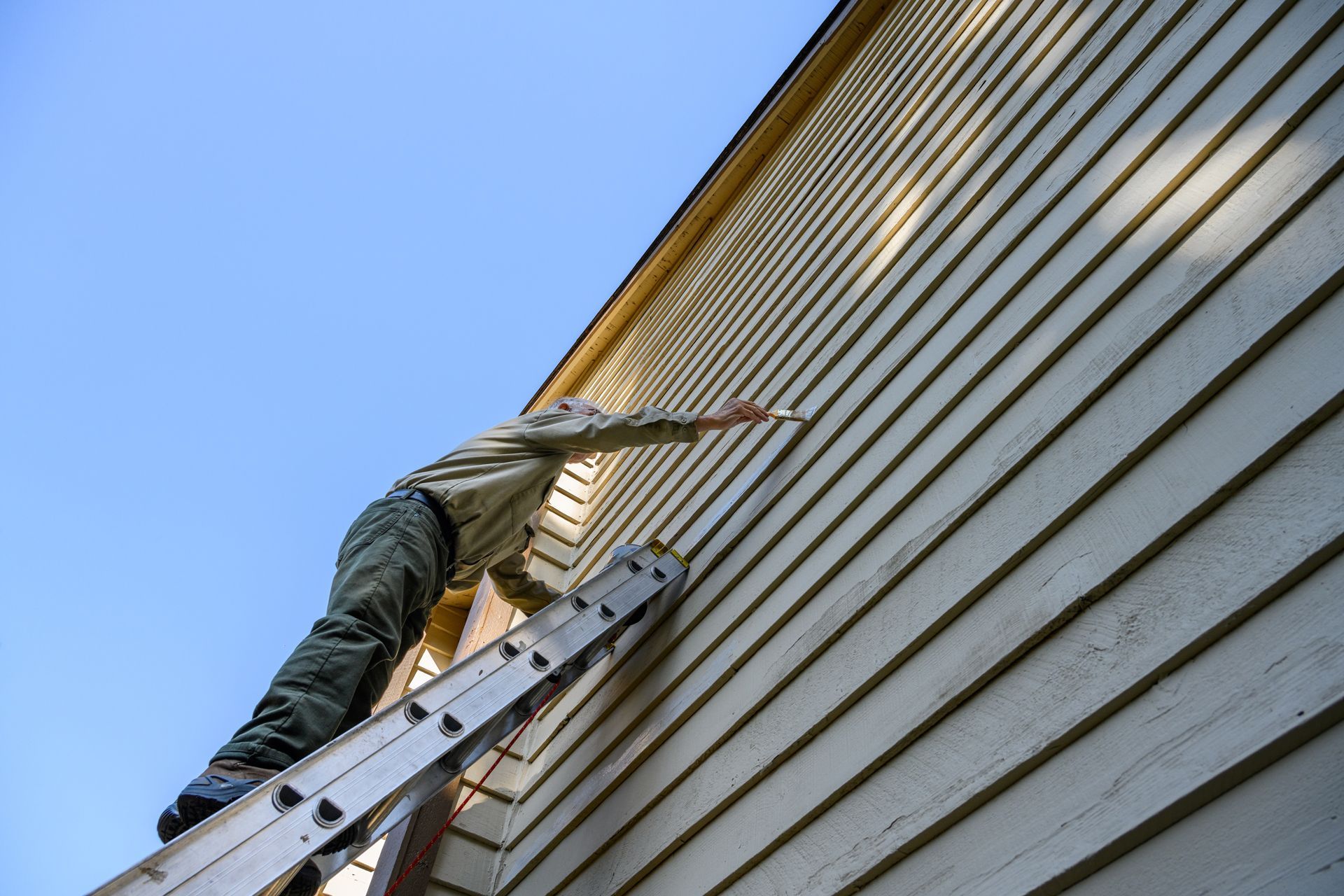 Person on a ladder painting the siding of a house, reaching upward against a blue sky.