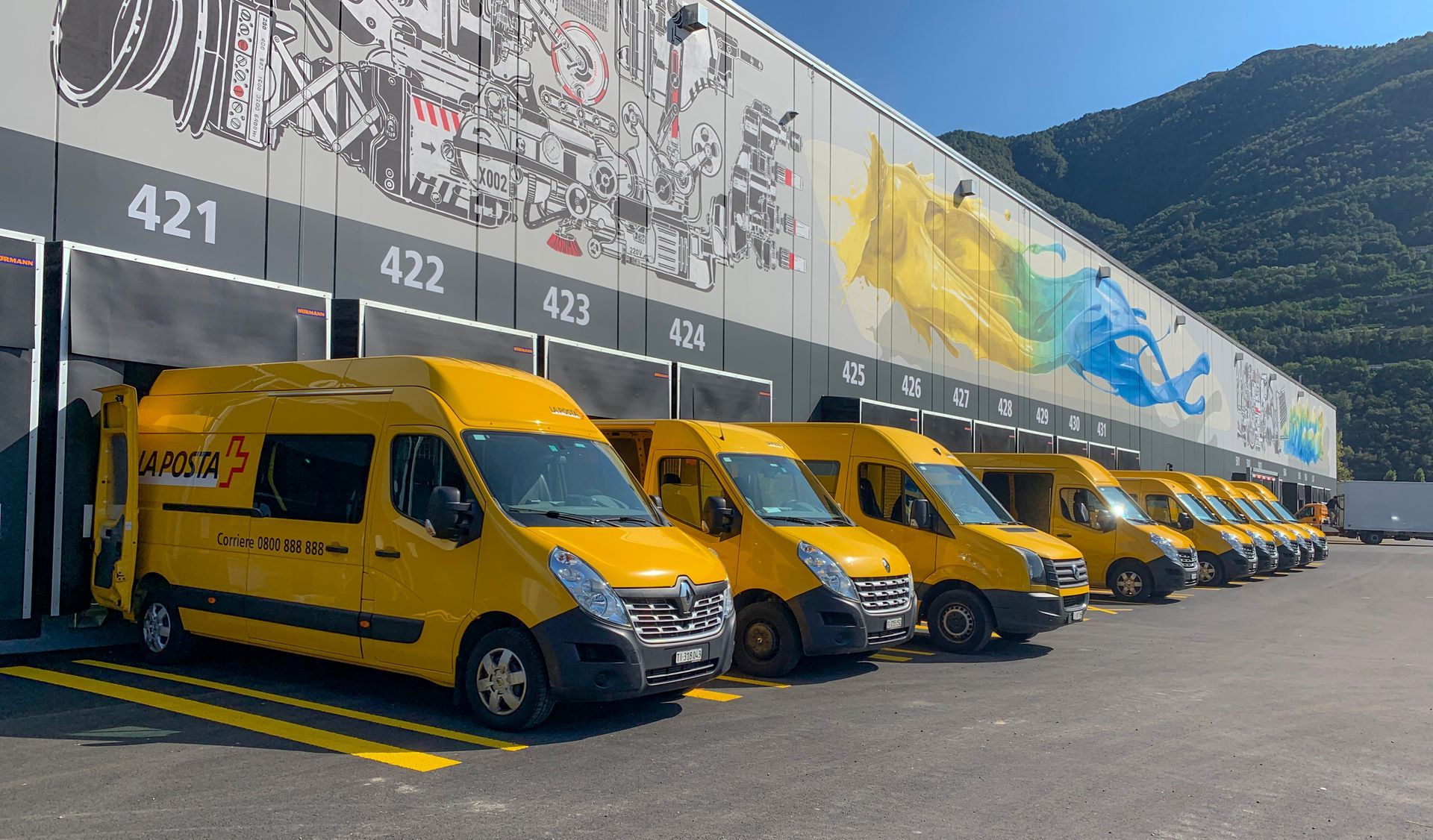 Furgoni gialli per le consegne in fila presso una banchina di carico. Sullo sfondo montagne, murale sull'edificio.