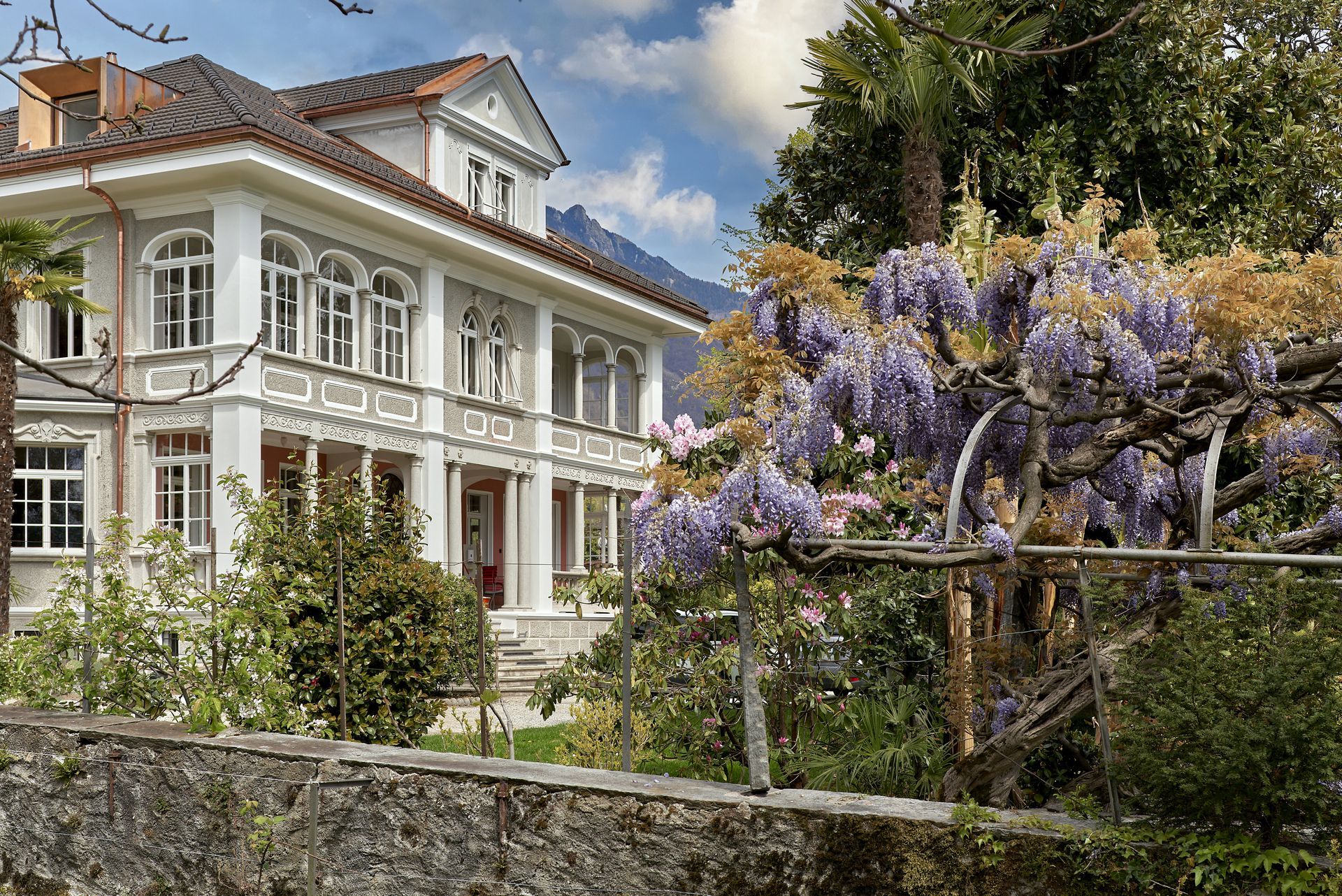 Villa bianca a due piani con balcone, circondata dal verde e da glicini viola in fiore.