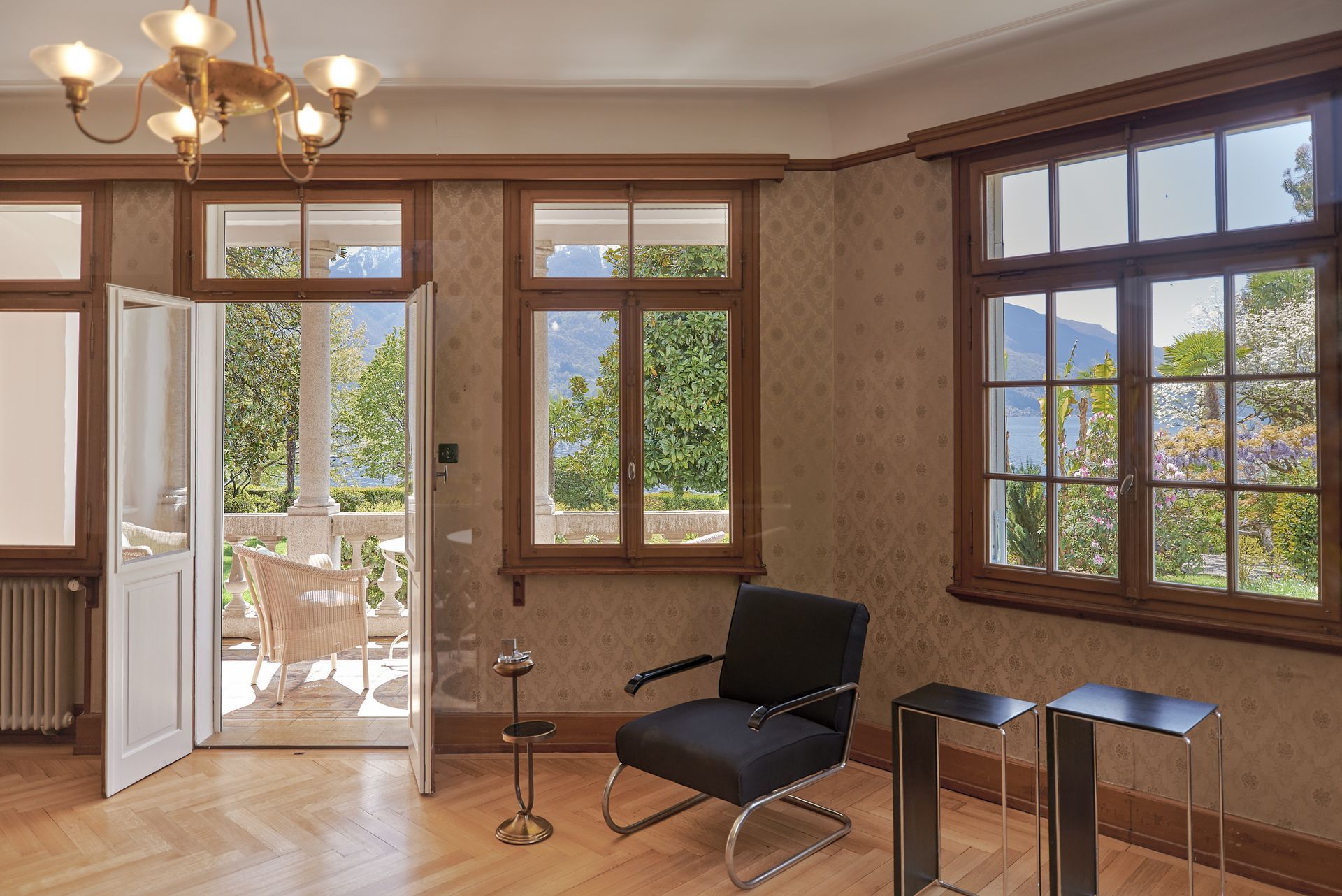 Camera con porte che danno su un balcone, finestre in legno, sedia nera e vista sugli alberi e lago in lontananza.