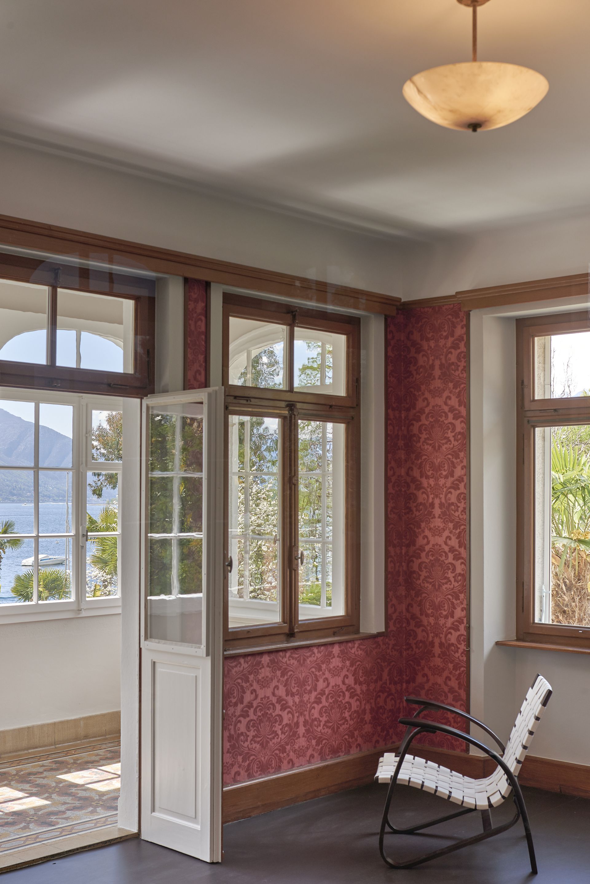 Stanza con pareti decorate con motivi rossi, finestre in legno con vista sull'acqua, sedia bianca e lampada.