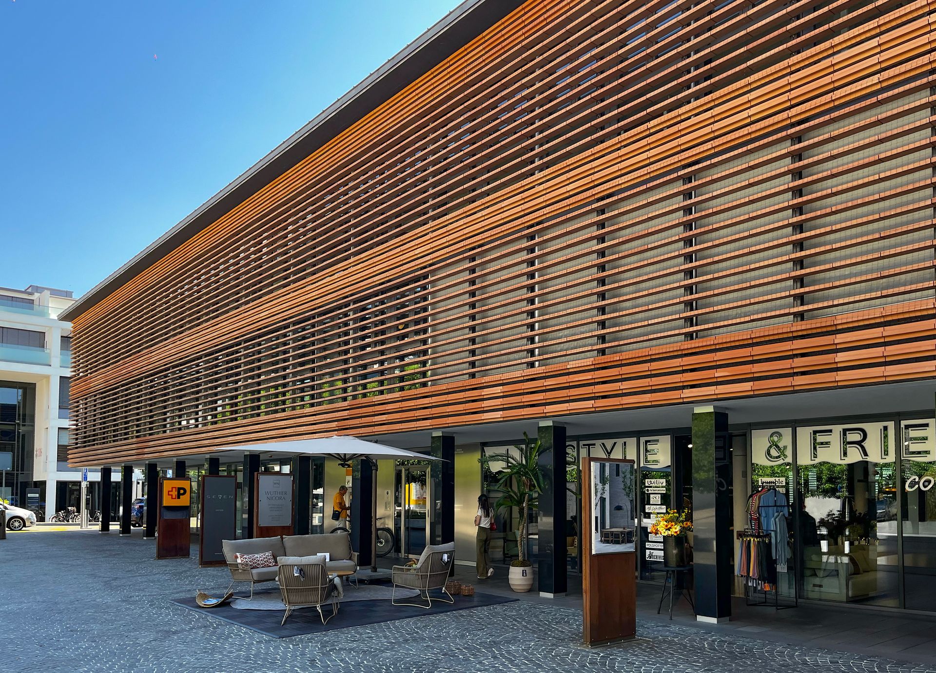 Edificio moderno con facciata marrone, tetto di tegole e posti a sedere all'aperto in un ambiente soleggiato.