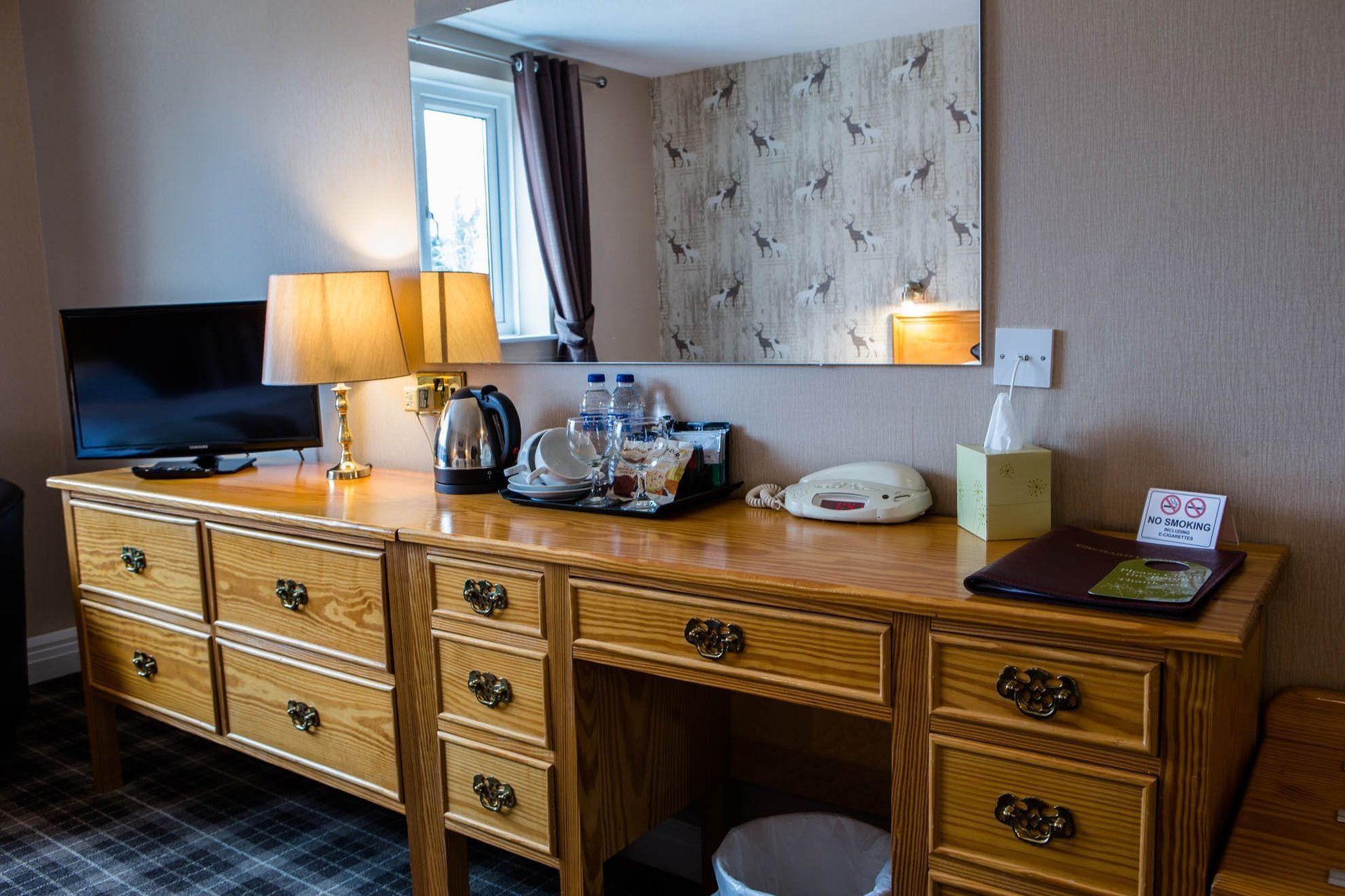 A hotel room with a desk , dresser , television and mirror.