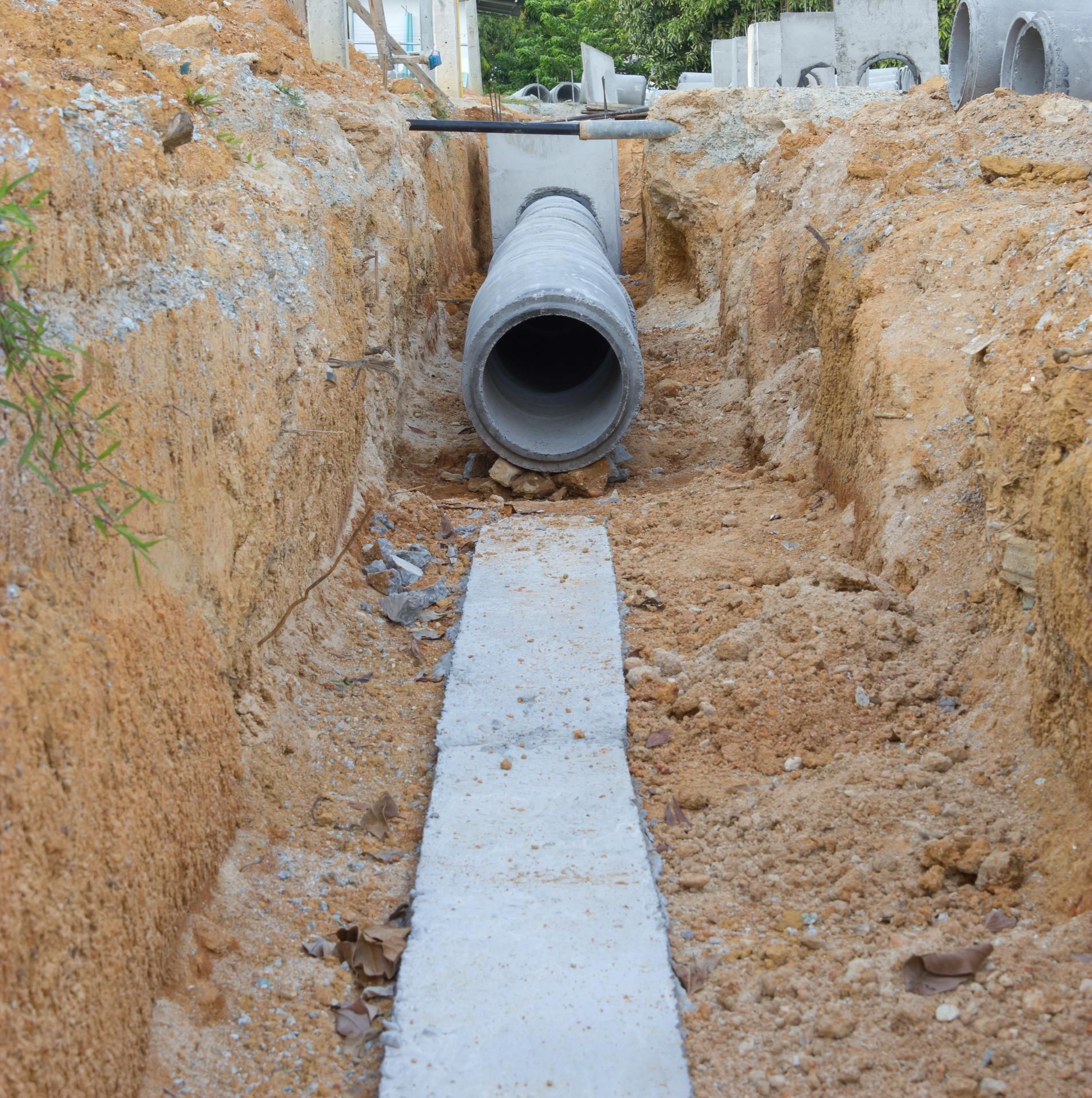 A concrete drainage pipe is installed at the end of a trench lined with a long concrete foundation base.
