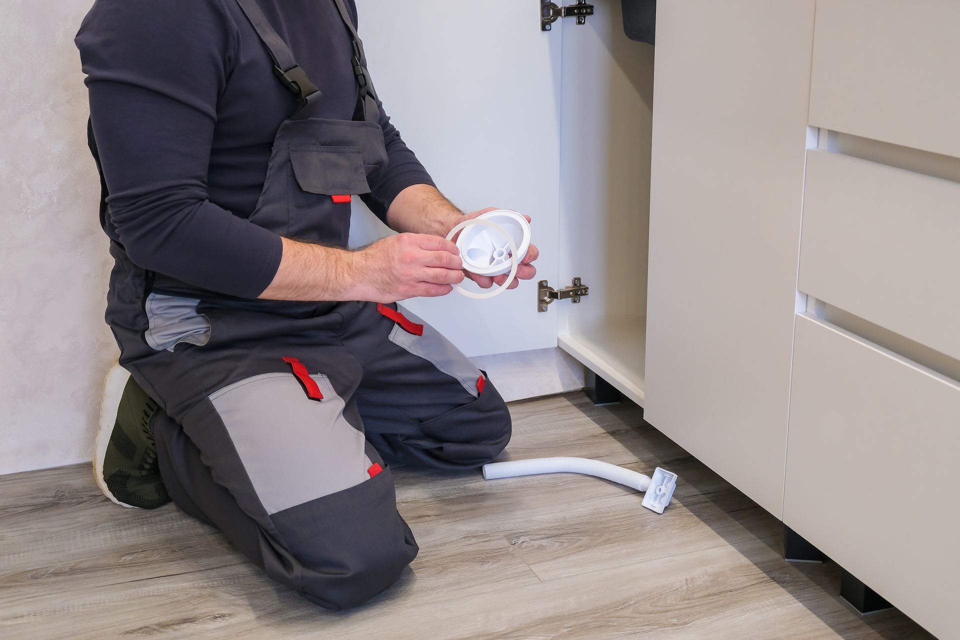 Technician kneeling by cabinet installing under-sink plumbing part and drain hose. Technician kneeling by cabinet installing under-sink plumbing part and drain hose.
