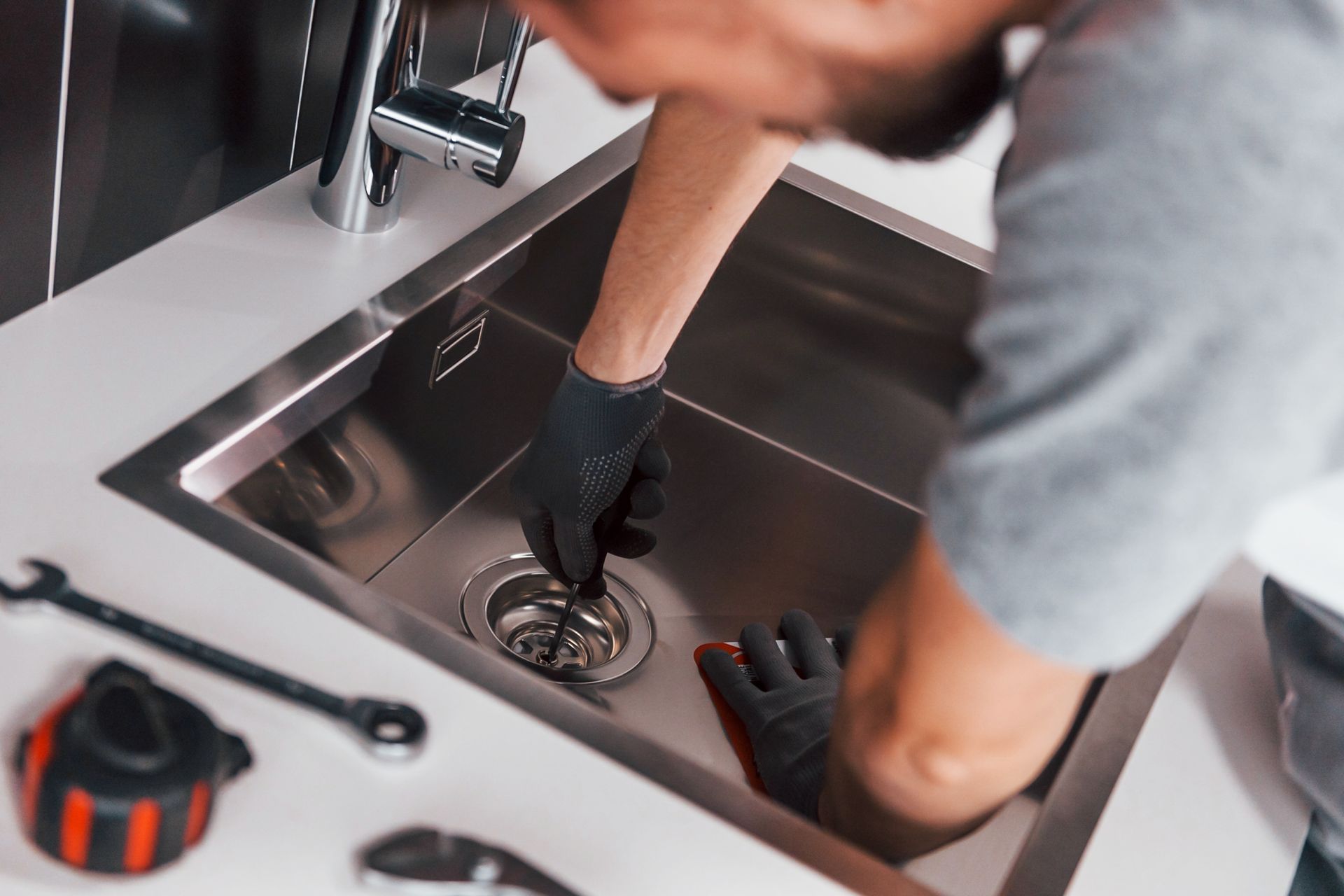 a person wearing black gloves using a tool to unclog a stainless steel kitchen sink drain.