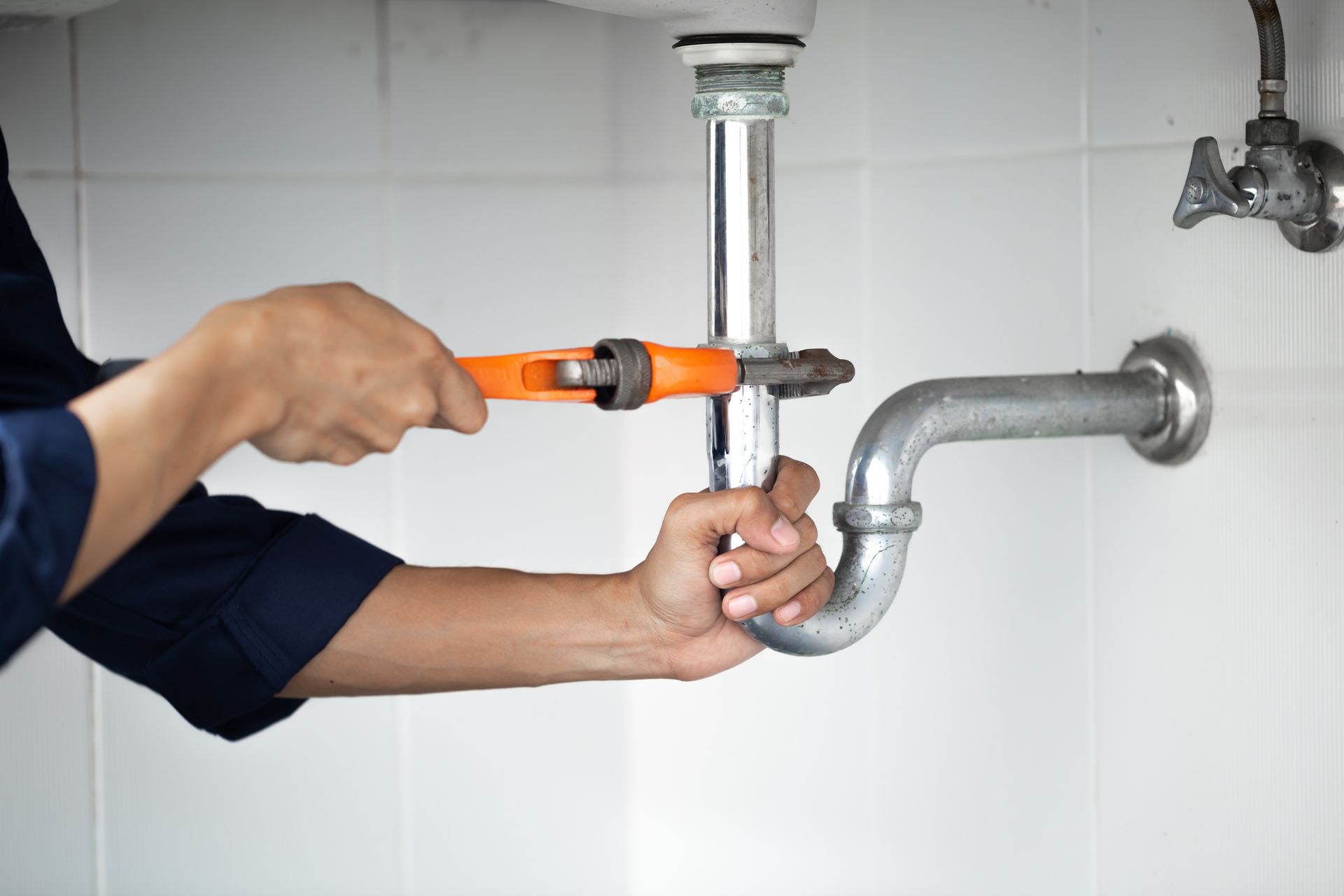 Plumber using an orange adjustable wrench on a chrome sink pipe under a white sink.