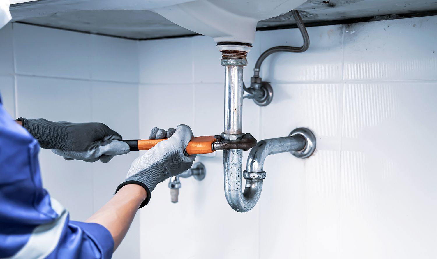 Plumber in blue uniform uses a wrench to tighten a pipe under a white sink. Plumber in blue uniform uses a wrench to tighten a pipe under a white sink.