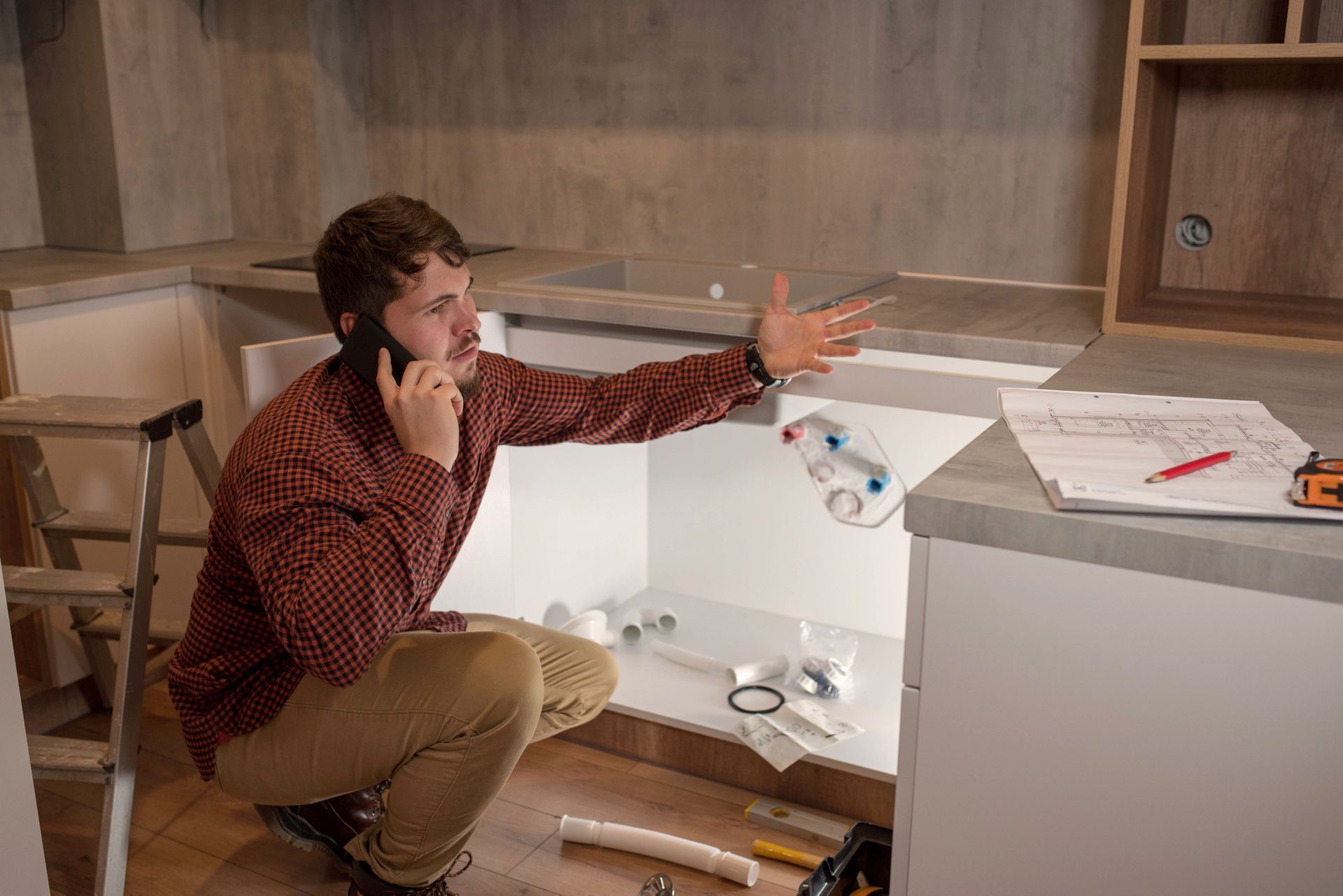 Man on phone fixing kitchen sink with tools and blueprint, kneeling near open cabinet. Man on phone fixing kitchen sink with tools and blueprint, kneeling near open cabinet.