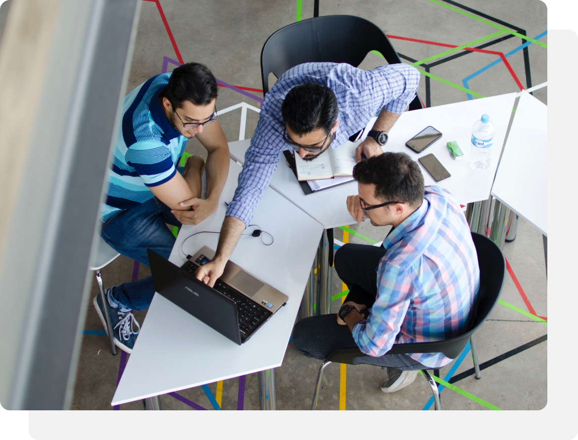 Three people around a white table, one using a laptop, looking at a notebook and phone, and talking in an office.