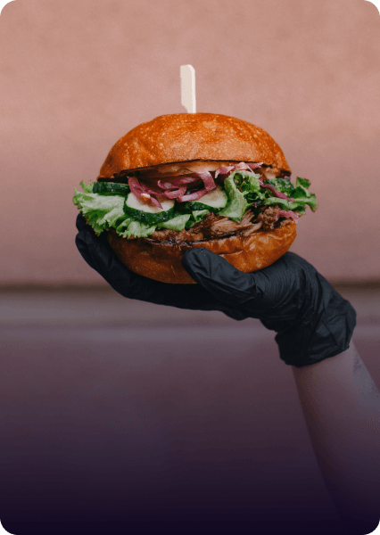 Hand holding a burger with lettuce, cucumbers, and onions on a toasted bun, held in a black glove.