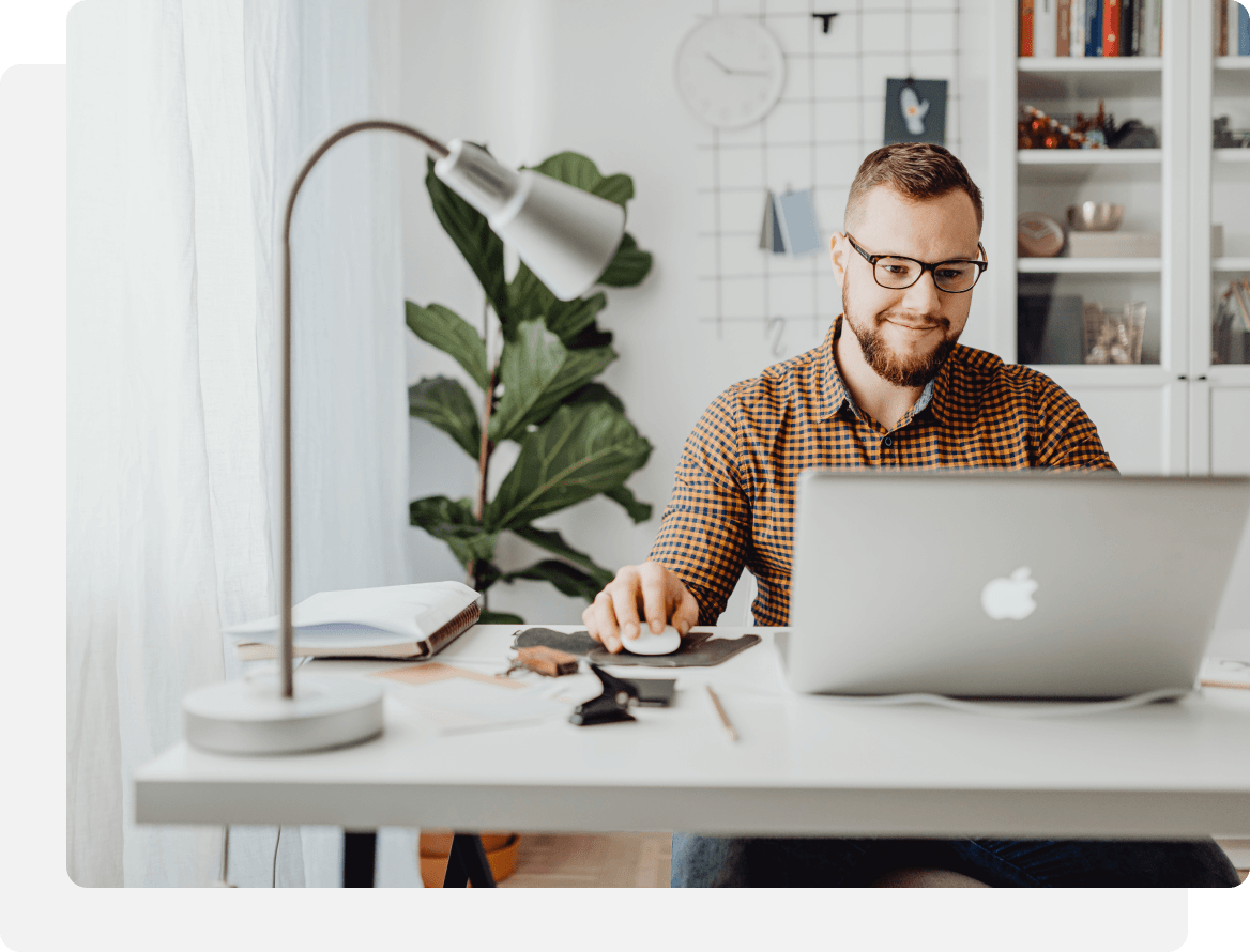 Man with glasses smiles, working on laptop at a desk in a bright room.