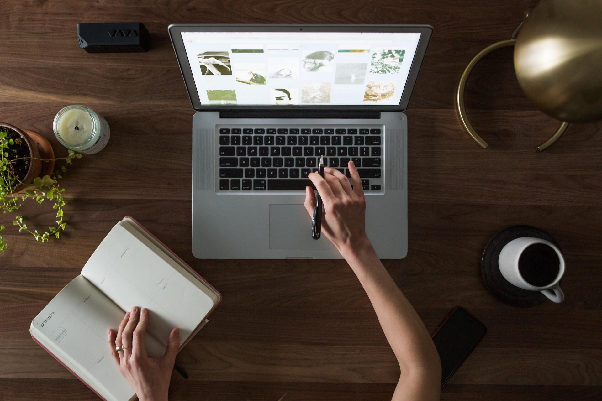 Laptop open on wooden desk, person typing, notebook, plant, candle, coffee cup, and phone.