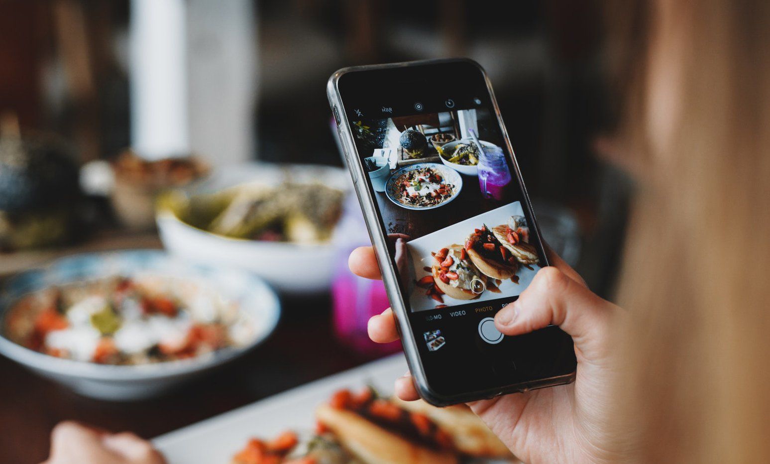 Person takes food photo with smartphone at restaurant table. Various dishes visible.