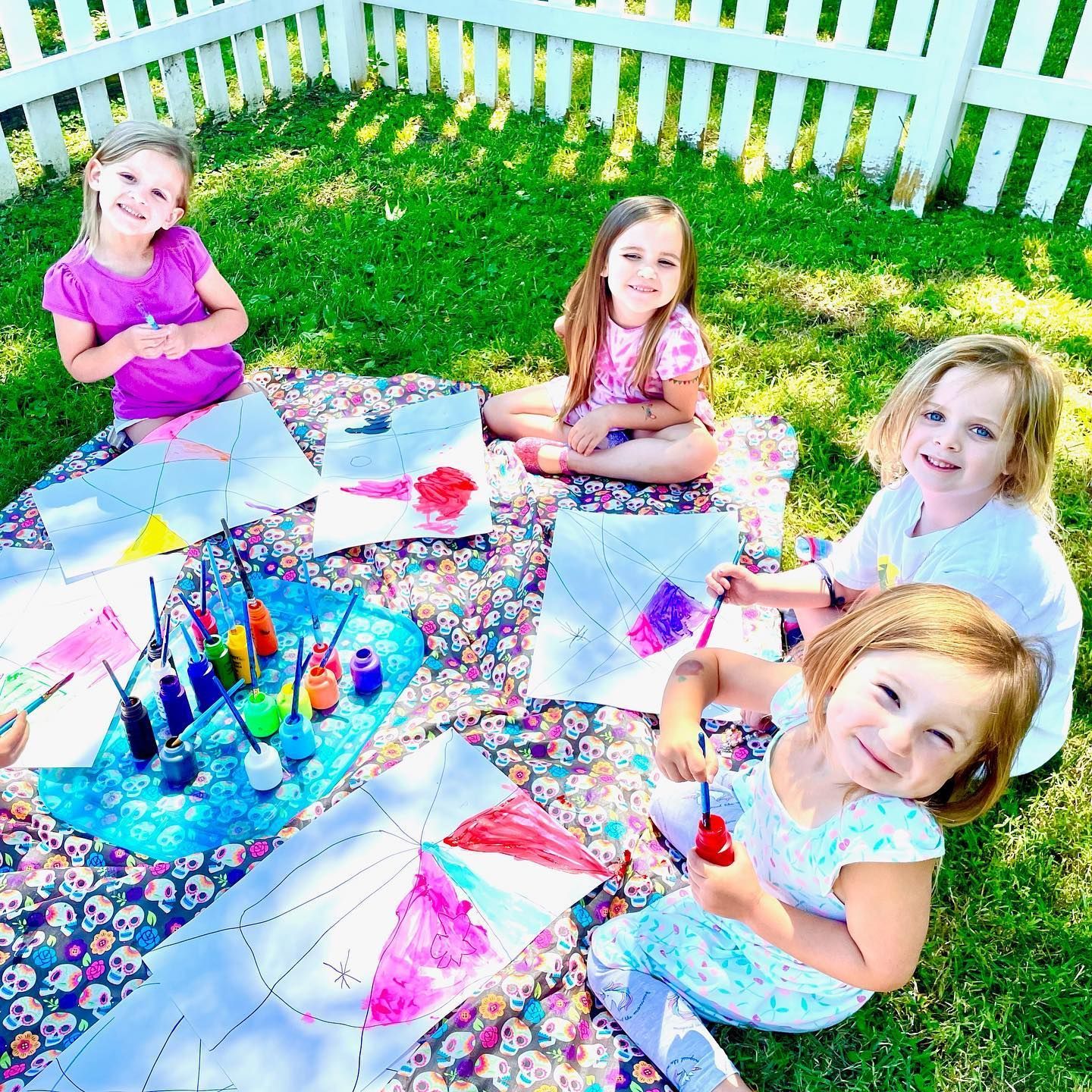 Five young girls painting colorful artwork on a blanket in a grassy yard near a white picket fence. They are smiling.