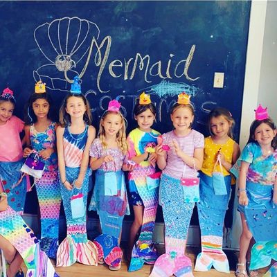 Group of young girls posing in front of a chalkboard decorated with a mermaid theme. They are wearing handmade mermaid tails and crowns.